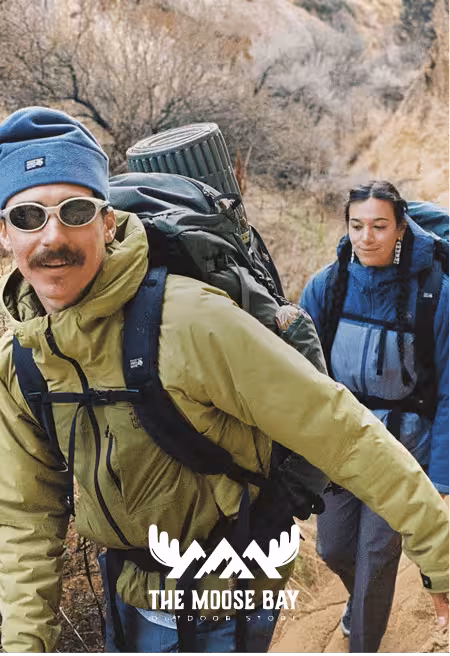 ITwo hikers wearing outdoor jackets and backpacks trekking on a nature trail, showcasing durable outdoor apparel and adventure gear by The Moose Bay Outdoor Shop in a rugged landscape.