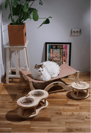 White fluffy cat resting on a handcrafted wooden cat hammock bed by WoWo Wood Wonder, with matching elevated wooden food and water bowl stands — eco-friendly and stylish pet furniture for modern homes.